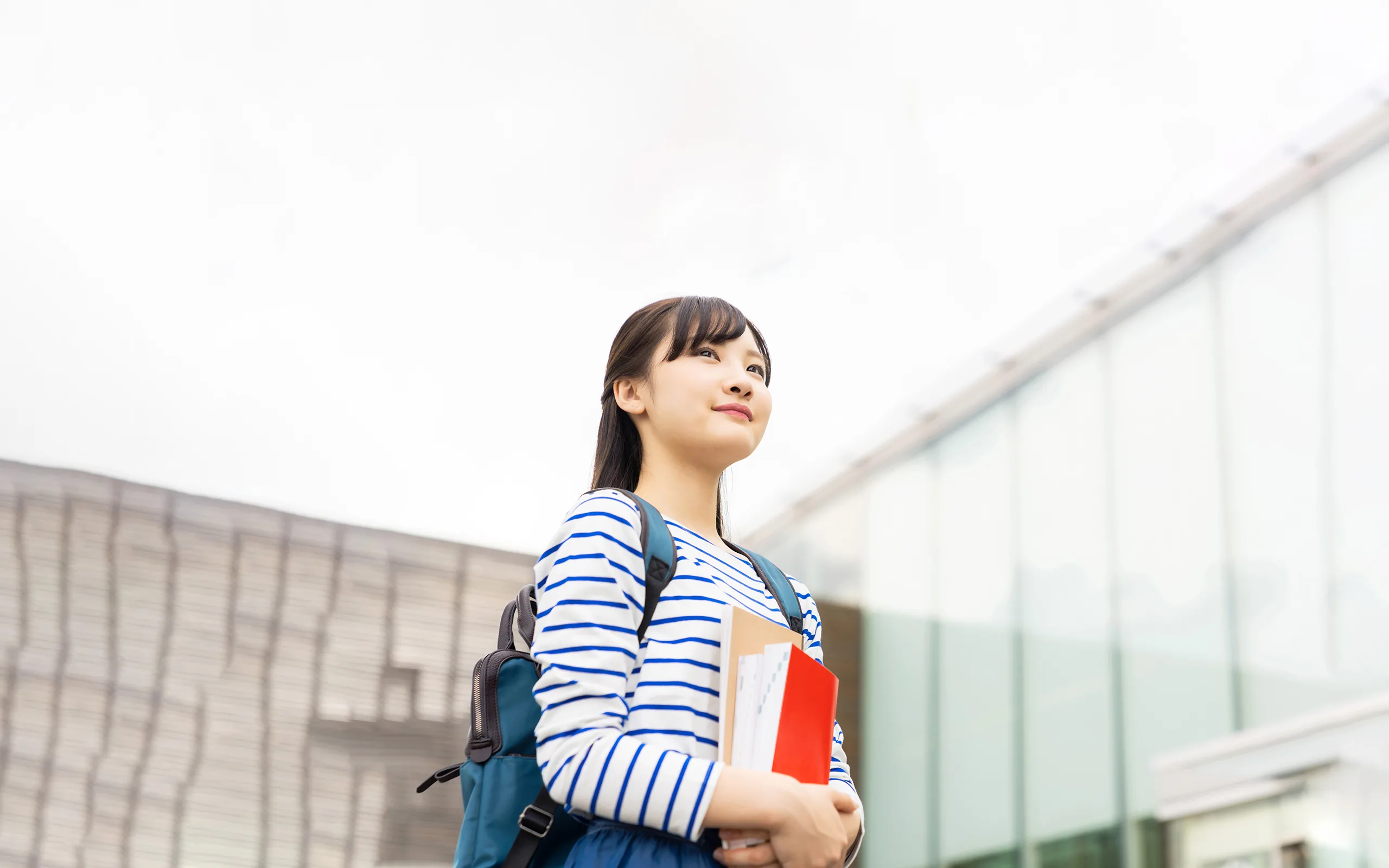 Photo d'une étudiante japonaise débout devant un bâtiment avec des grandes baies vitrées regardant au loin.